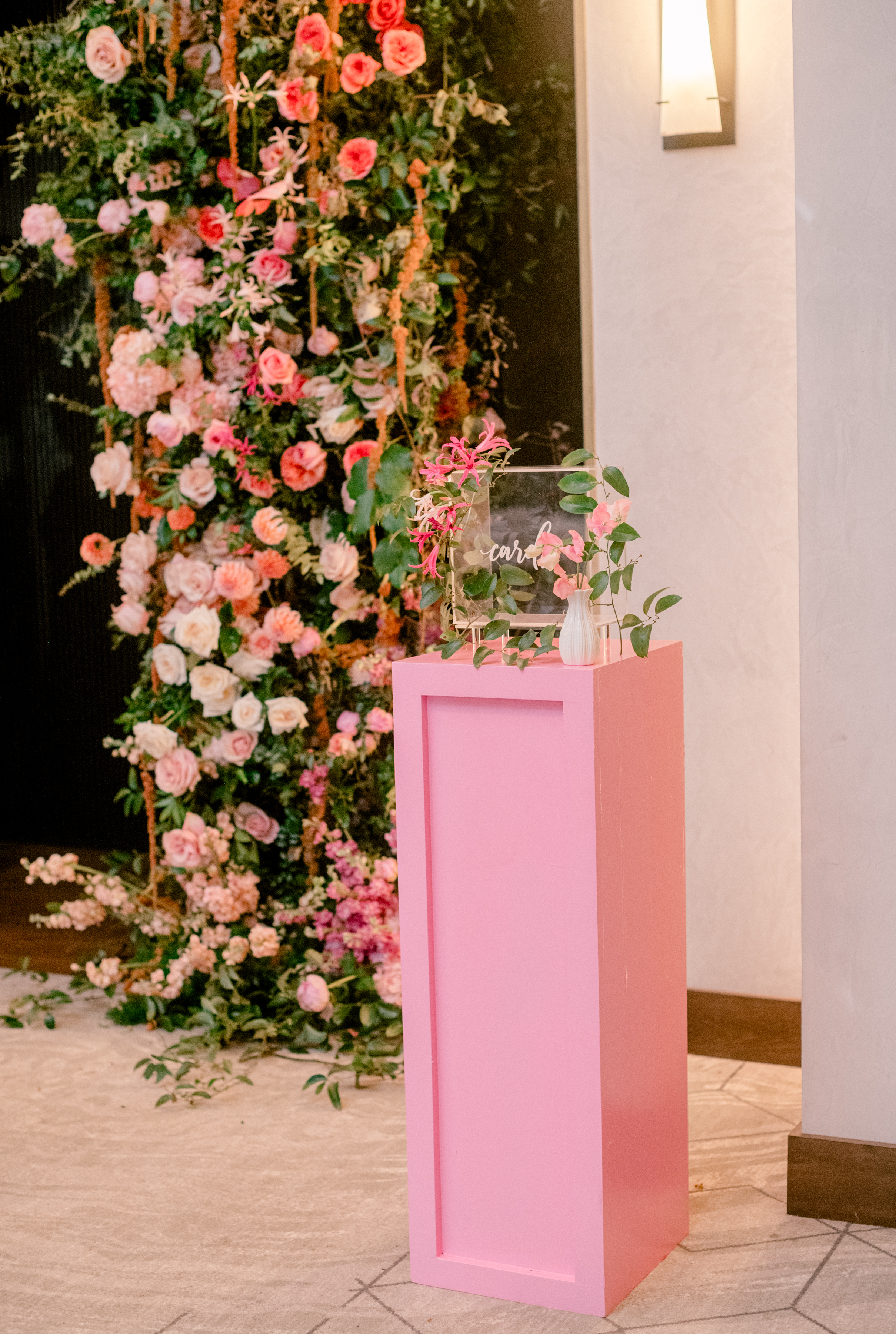 A pink pedestal with an acrylic card box and delicate floral accents stands beside a lush backdrop of pink and coral flowers.
