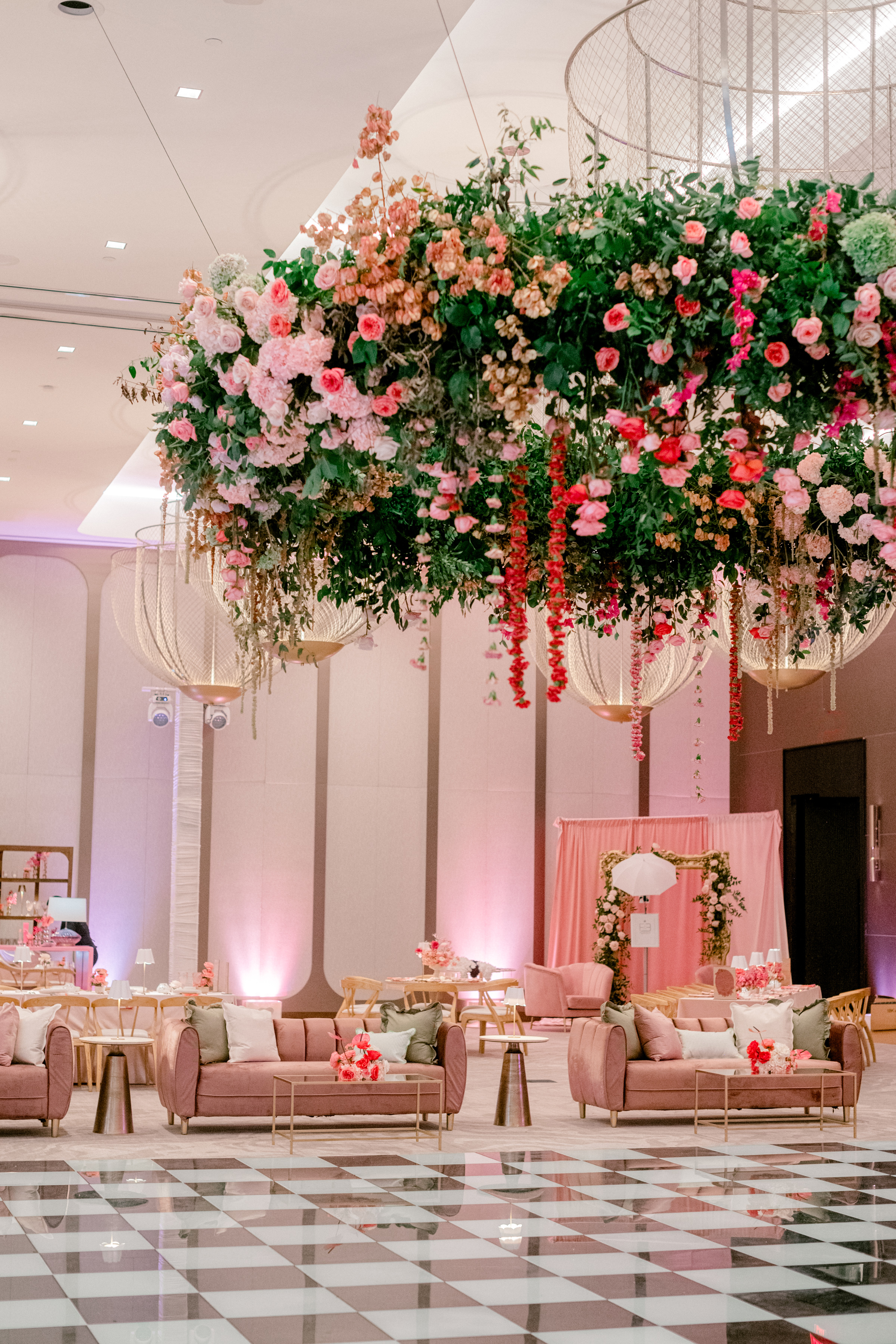 Suspended pink and coral floral installation hangs above a chic event lounge with velvet seating and a checkered dance floor.