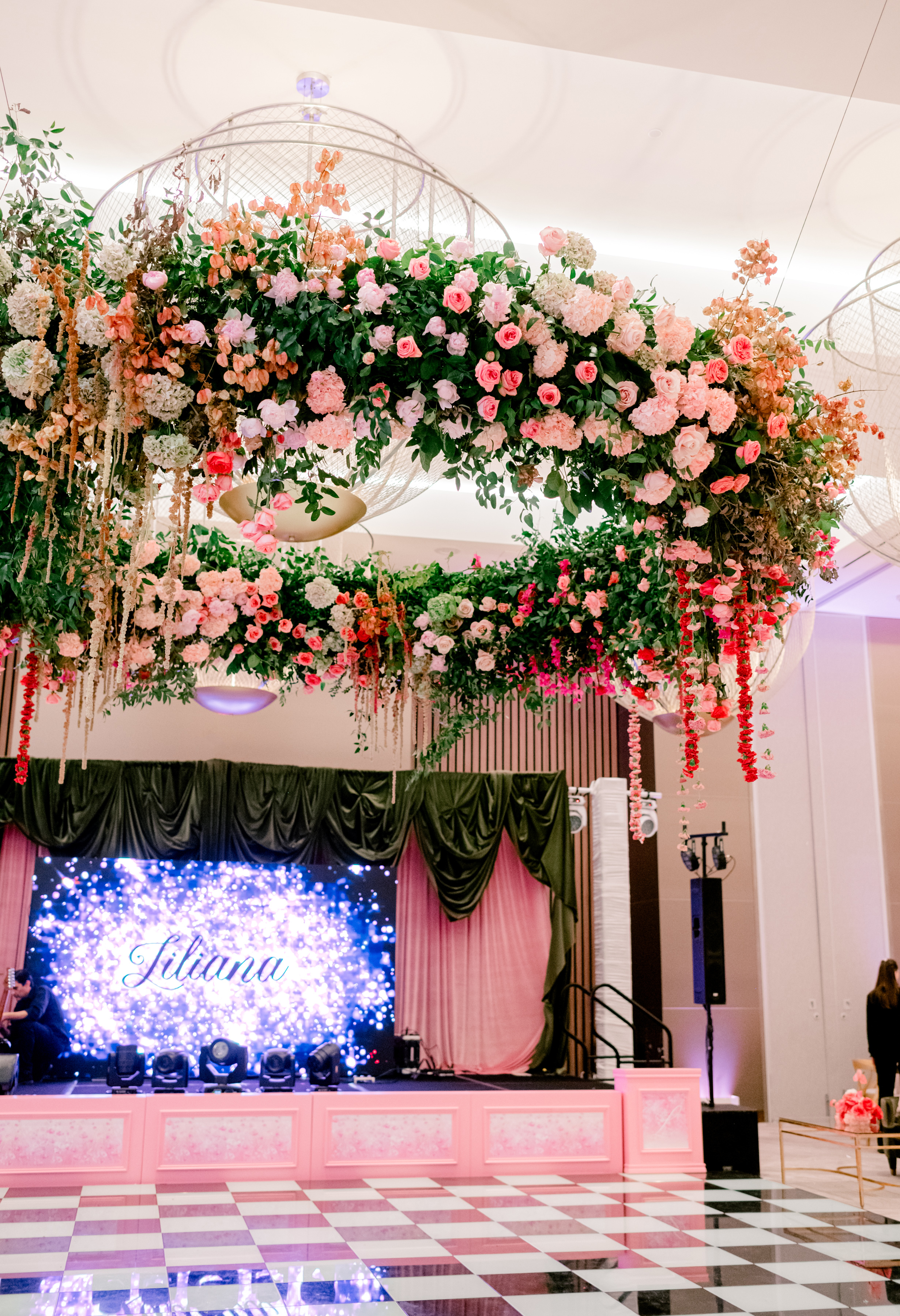 A suspended circular floral canopy of pink roses, hydrangeas, and trailing greenery hangs above a ballroom stage and checkered floor.
