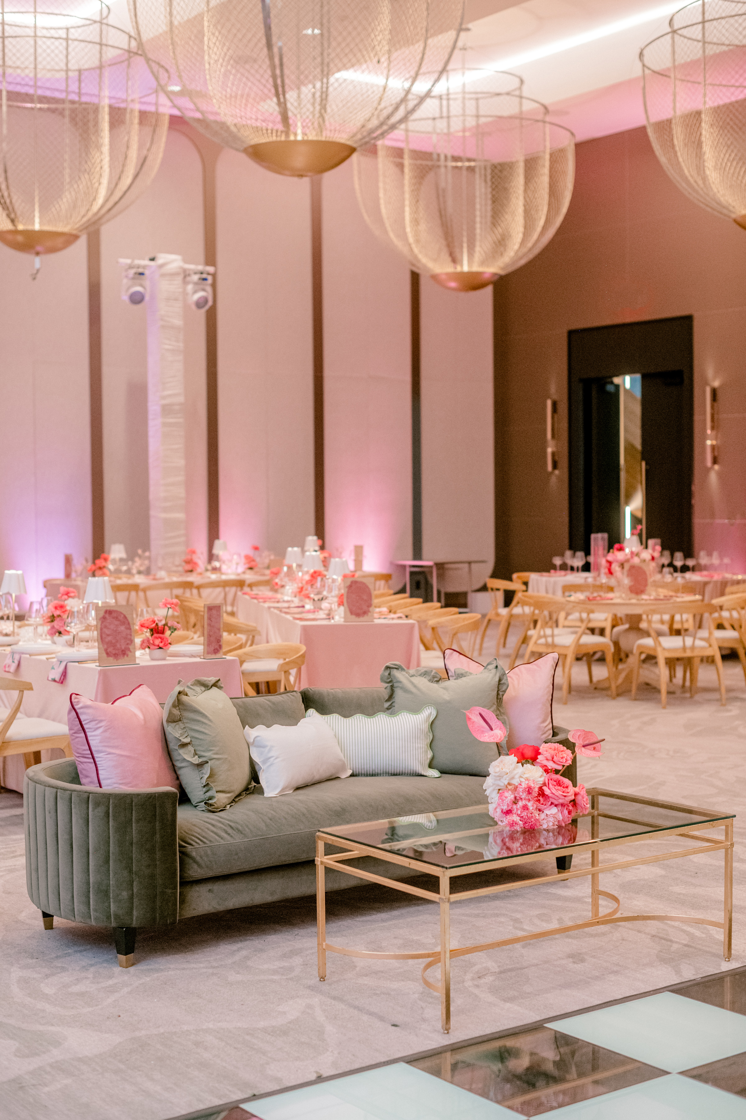 Sage green velvet lounge sofa with pastel pillows set before pink dining tables under sculpted wire chandeliers.