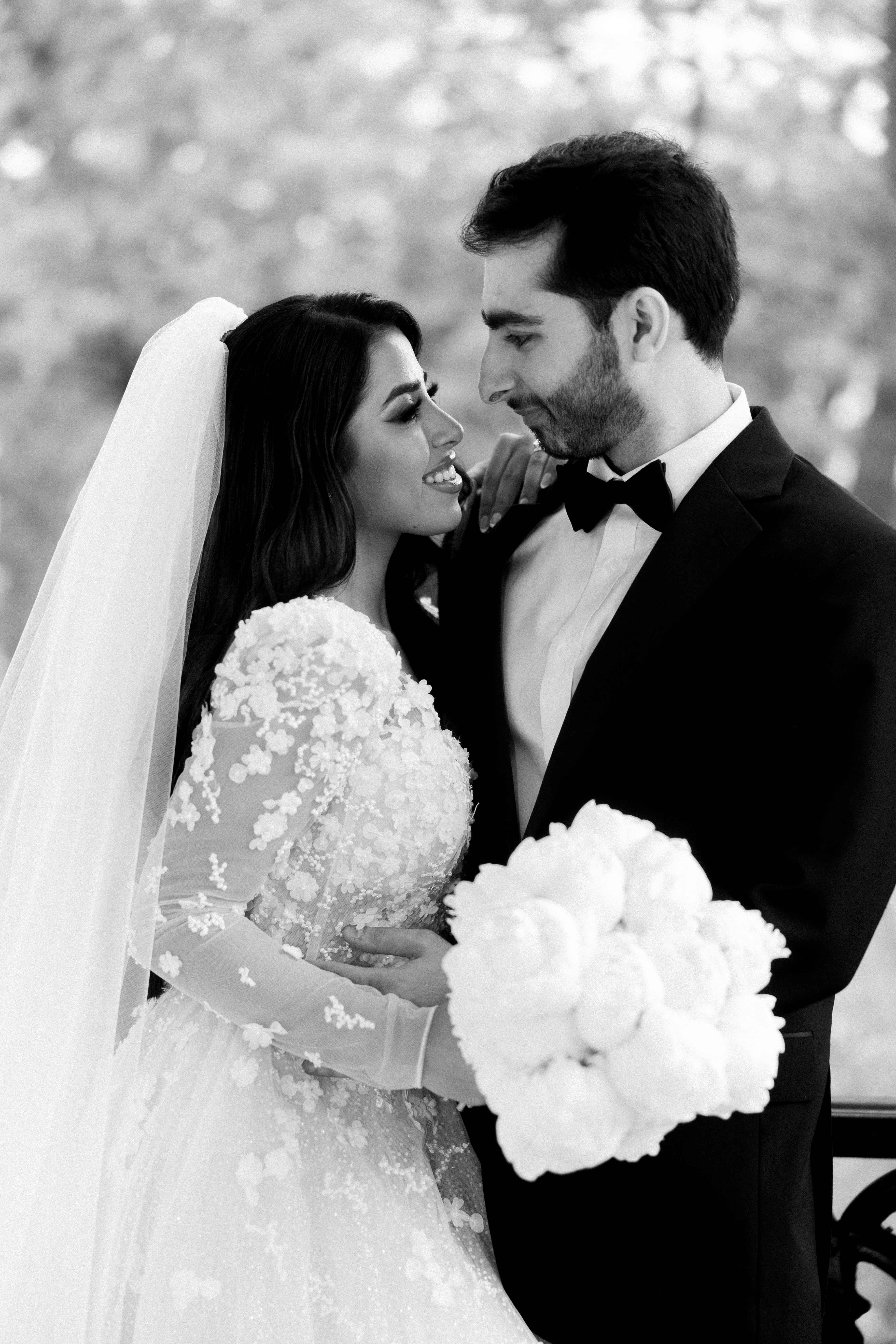 A smiling bride in an embroidered lace gown and groom in a classic tuxedo embracing outdoors.