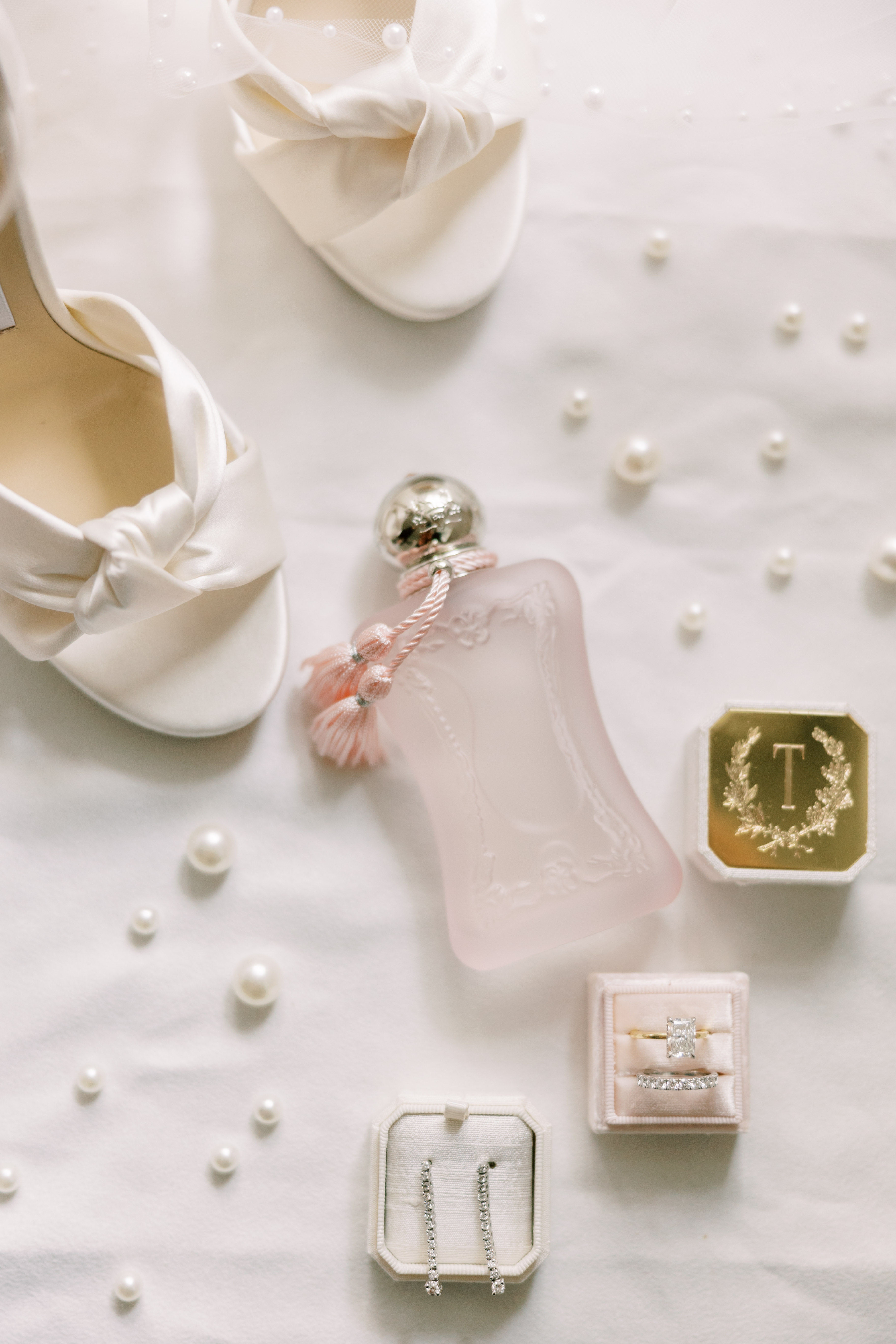 Bridal accessories including a blush pink perfume bottle, white heel, and diamond drop earrings displayed on soft fabric.