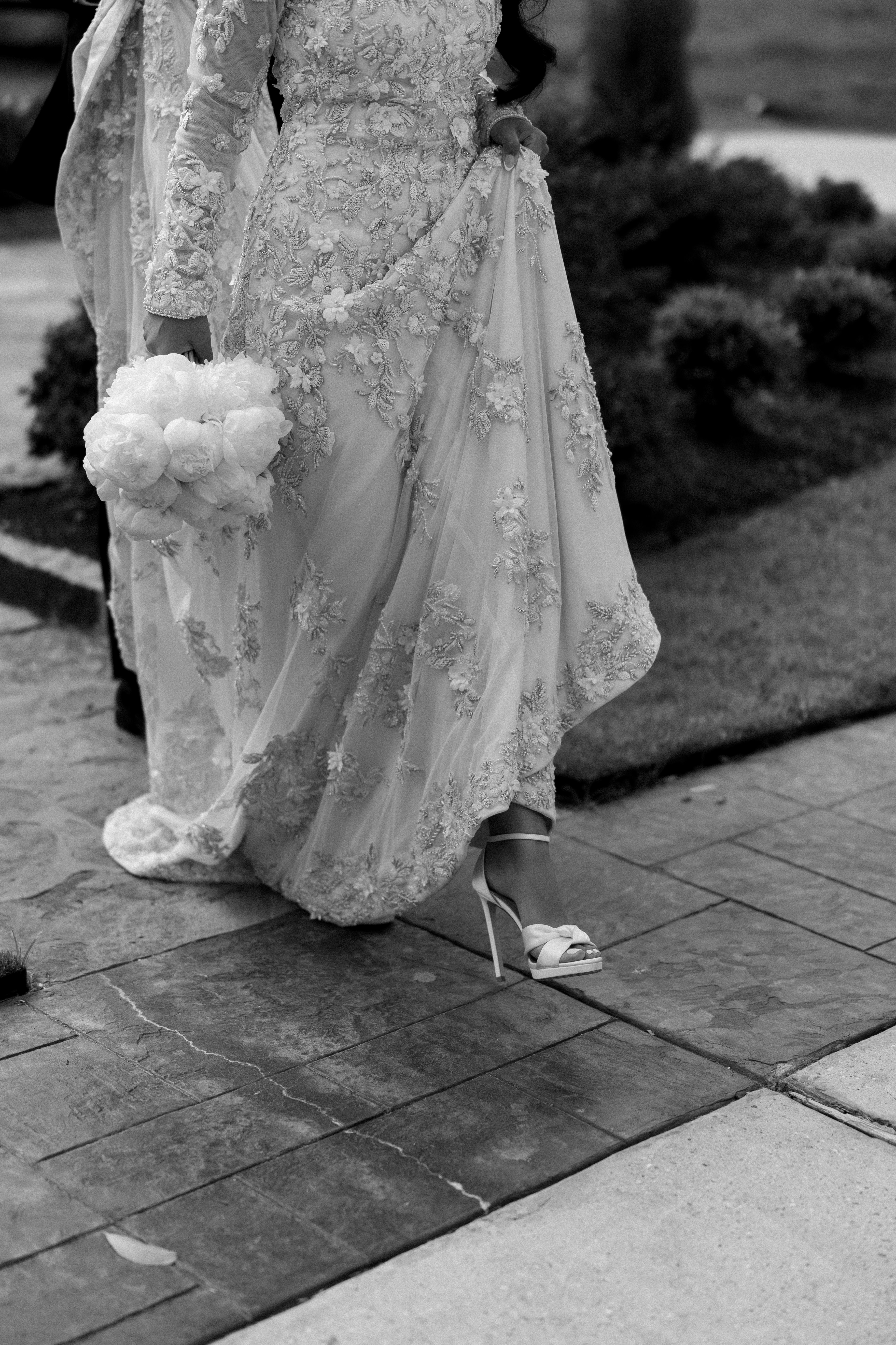 A bride lifts her intricately beaded floral embroidered wedding gown, revealing a white stiletto heel on a stone walkway.