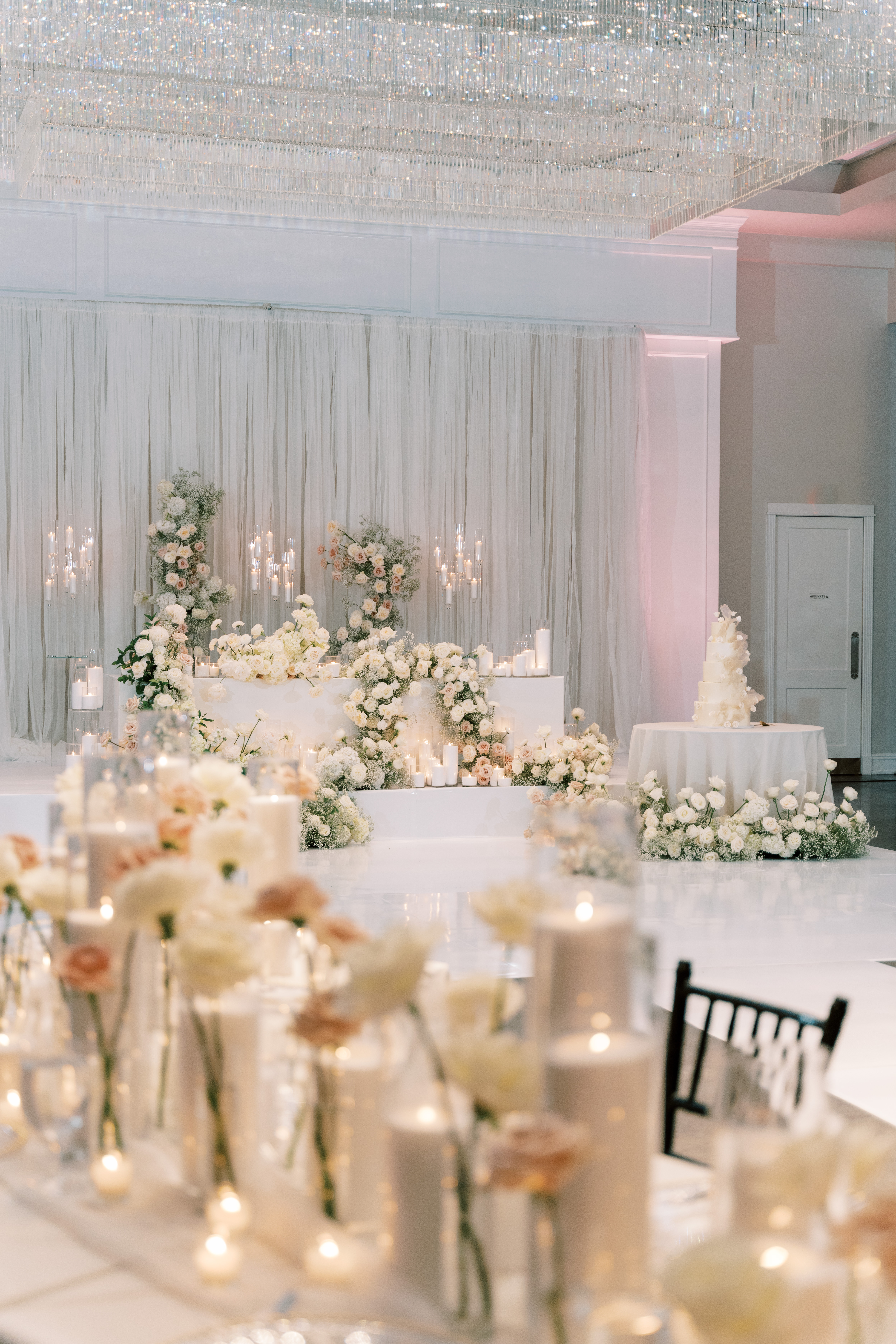 White wedding stage adorned with lush ivory roses, baby's breath, glowing pillar candles, and a grand crystal chandelier overhead.