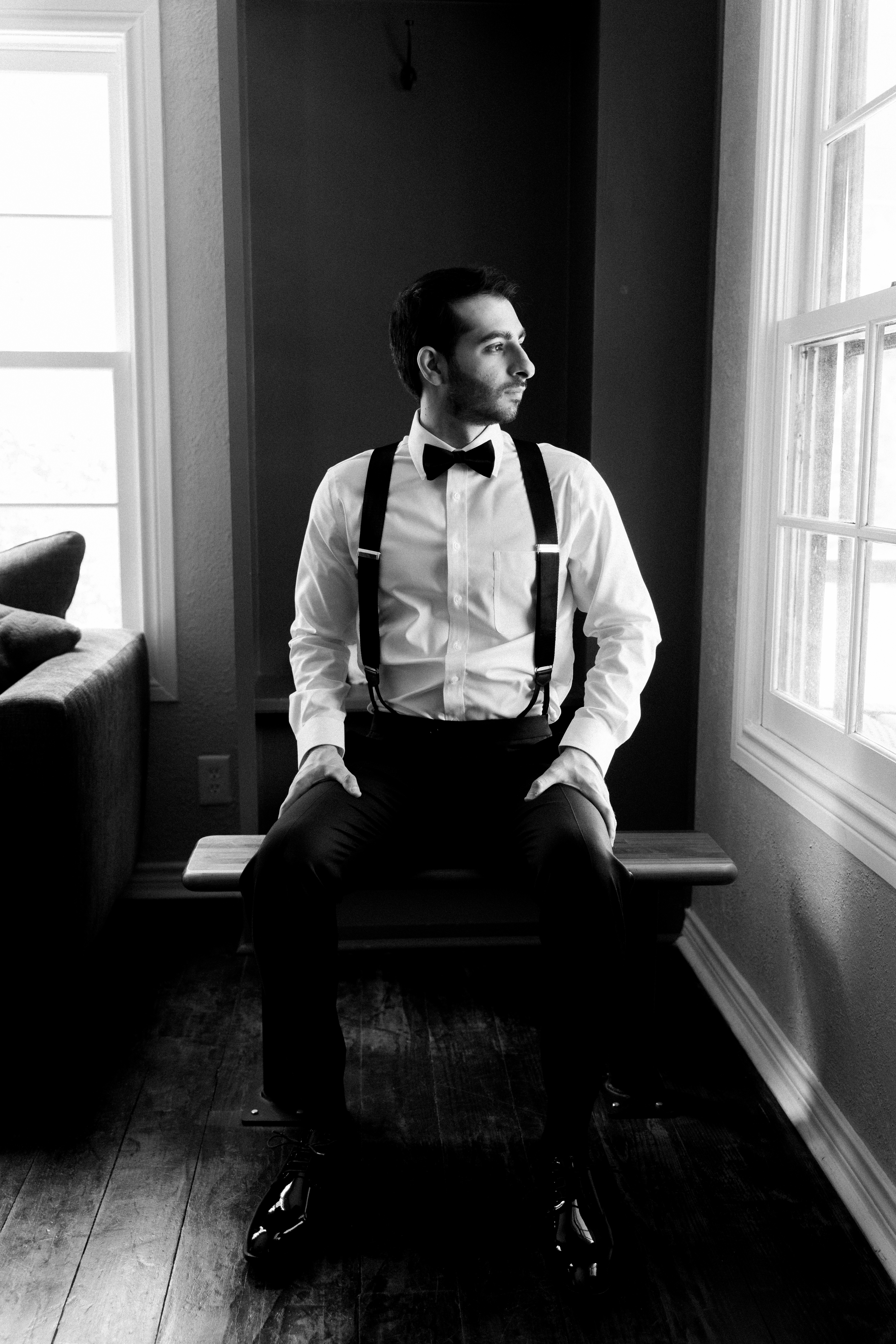 Groom in a white dress shirt, black bow tie, and suspenders seated in black and white.