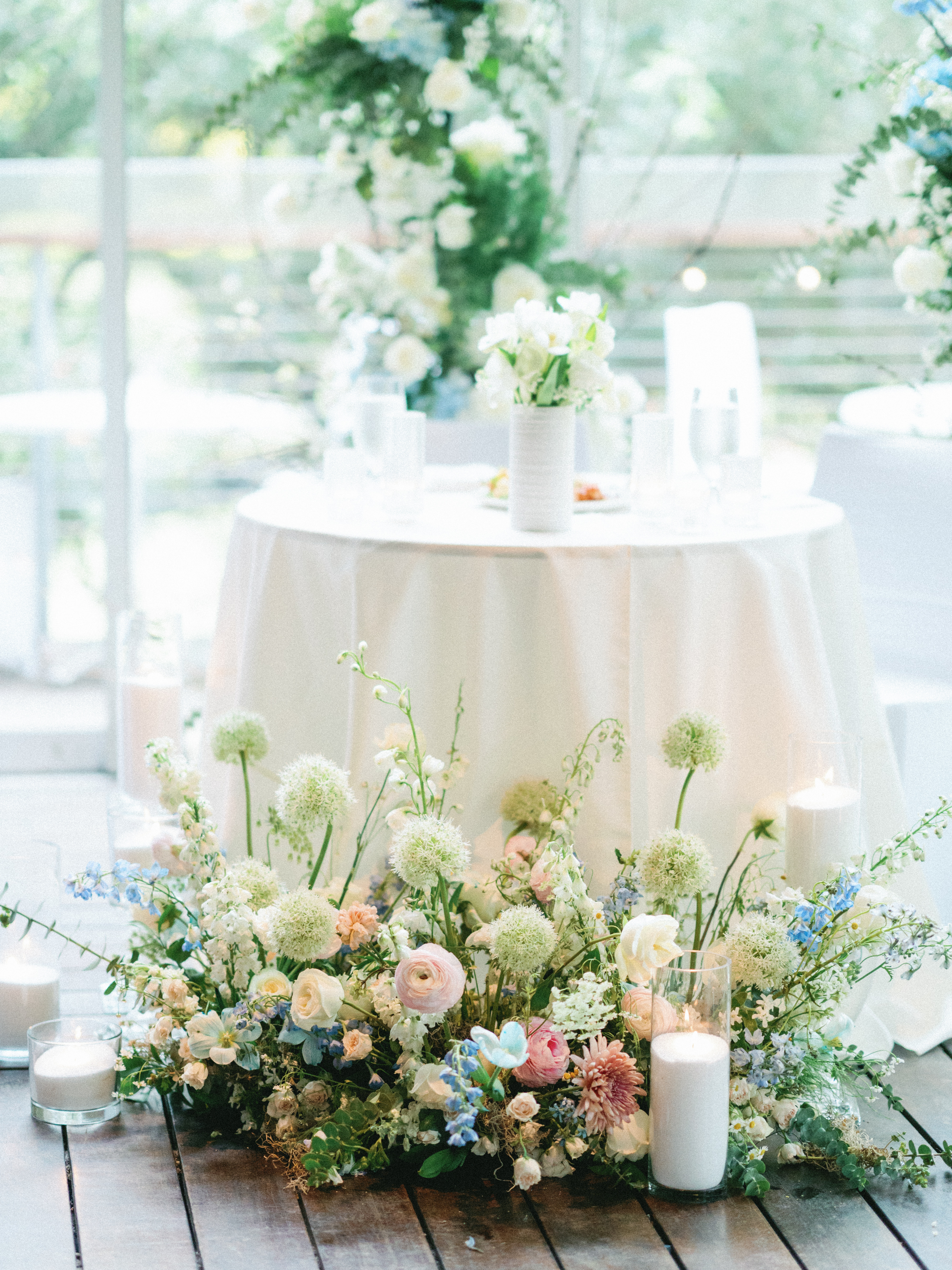 Lush floor florals with blush ranunculus and soft blue blossoms set before a white draped dining table with white flowers.