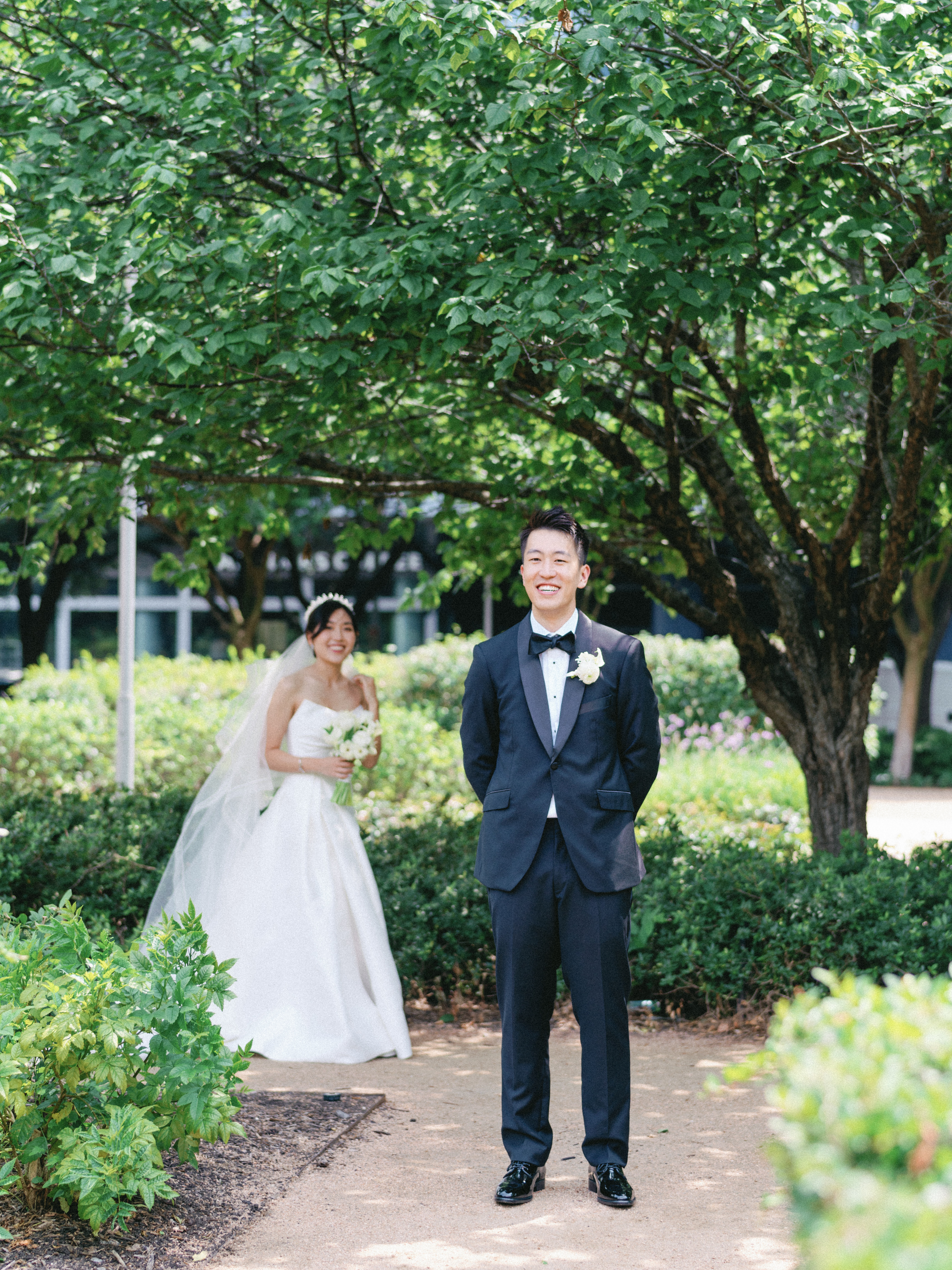 A smiling groom in a black tuxedo stands before an outdoor first look with the bride holding a white bouquet behind him.