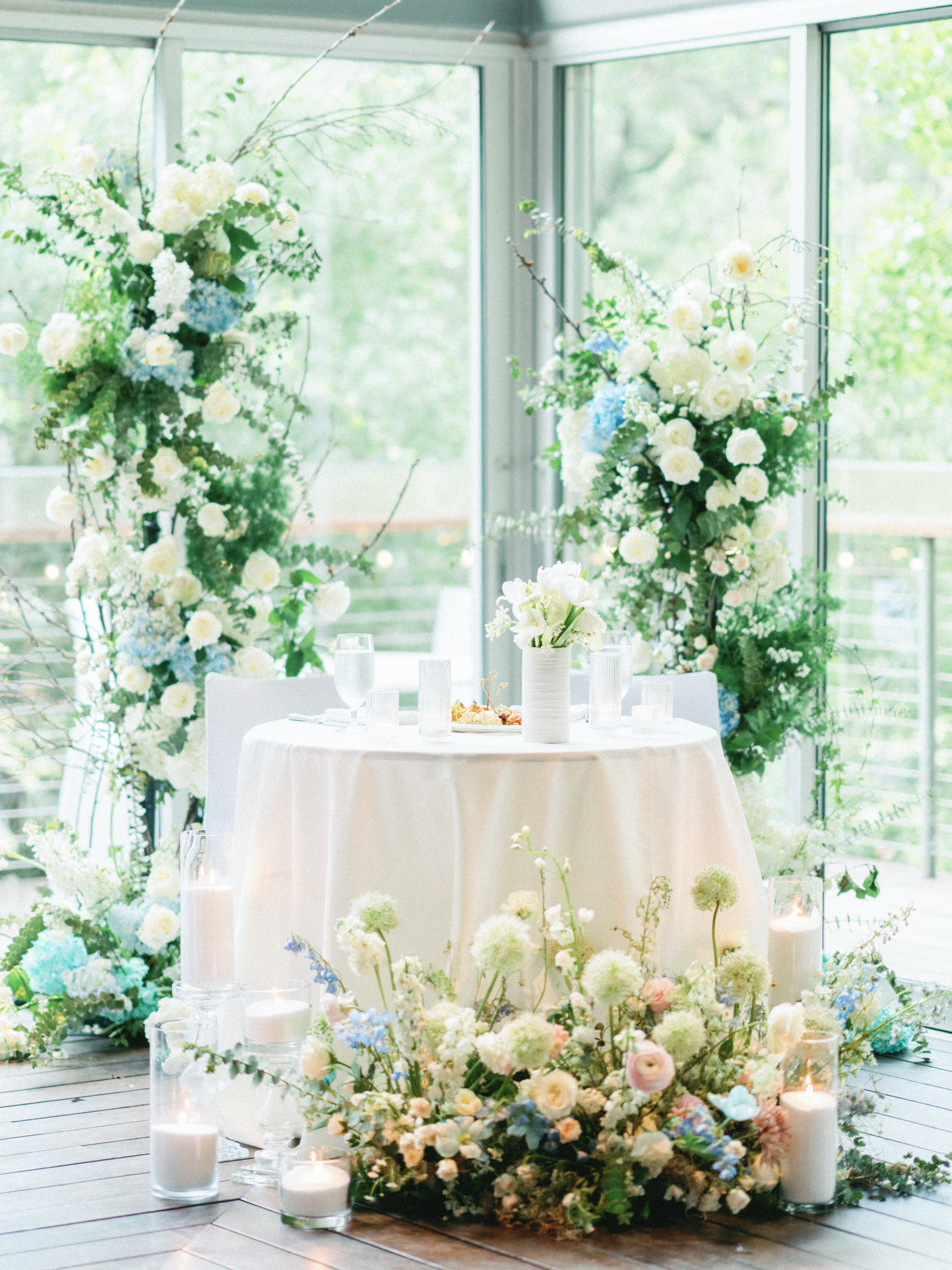 A wedding table set with white linens, fluted glassware, and lush floral arrangements featuring white, cream, and soft blue blooms.