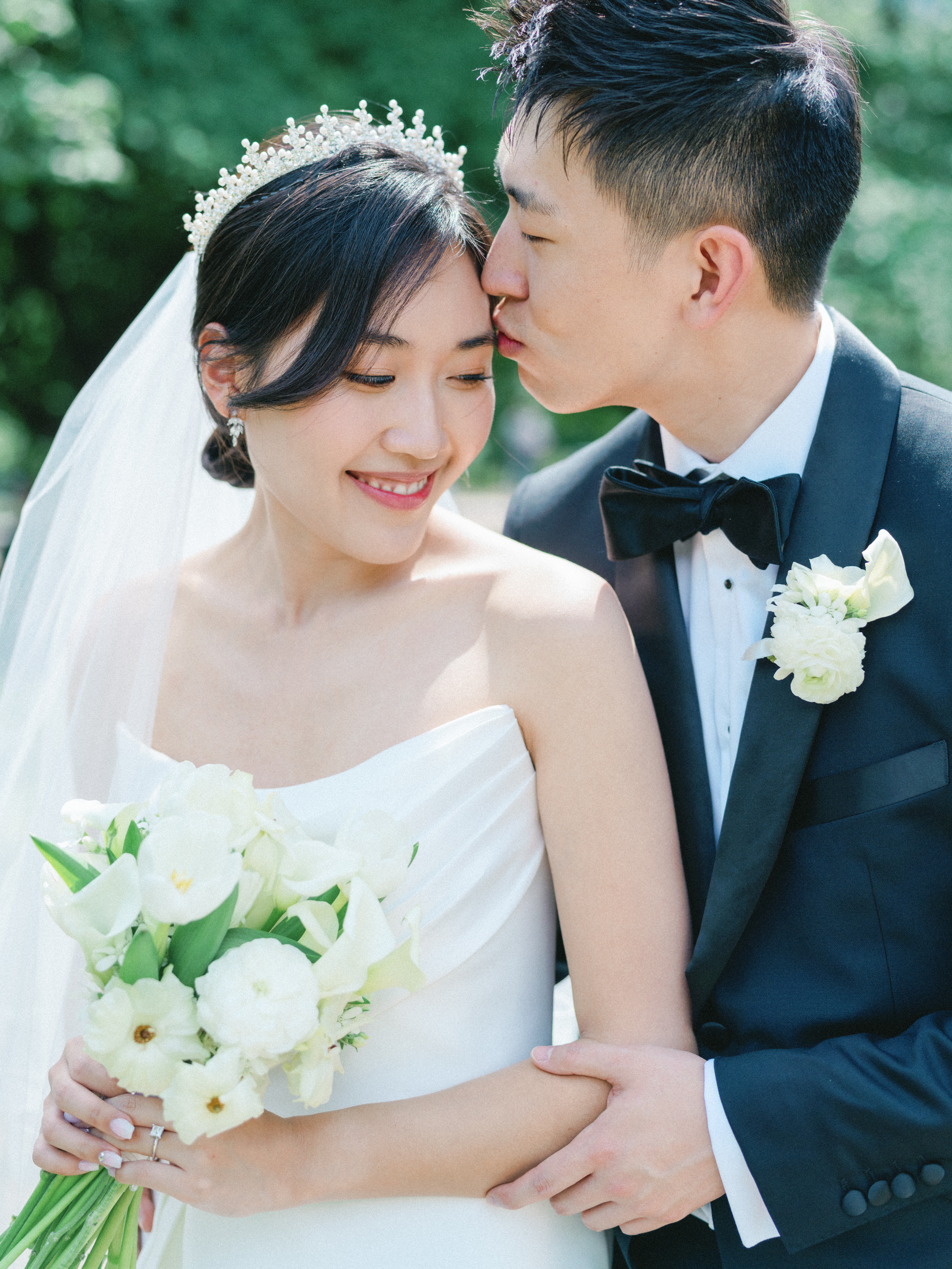 A groom gently kisses the bride's temple as she smiles during an outdoor wedding portrait.