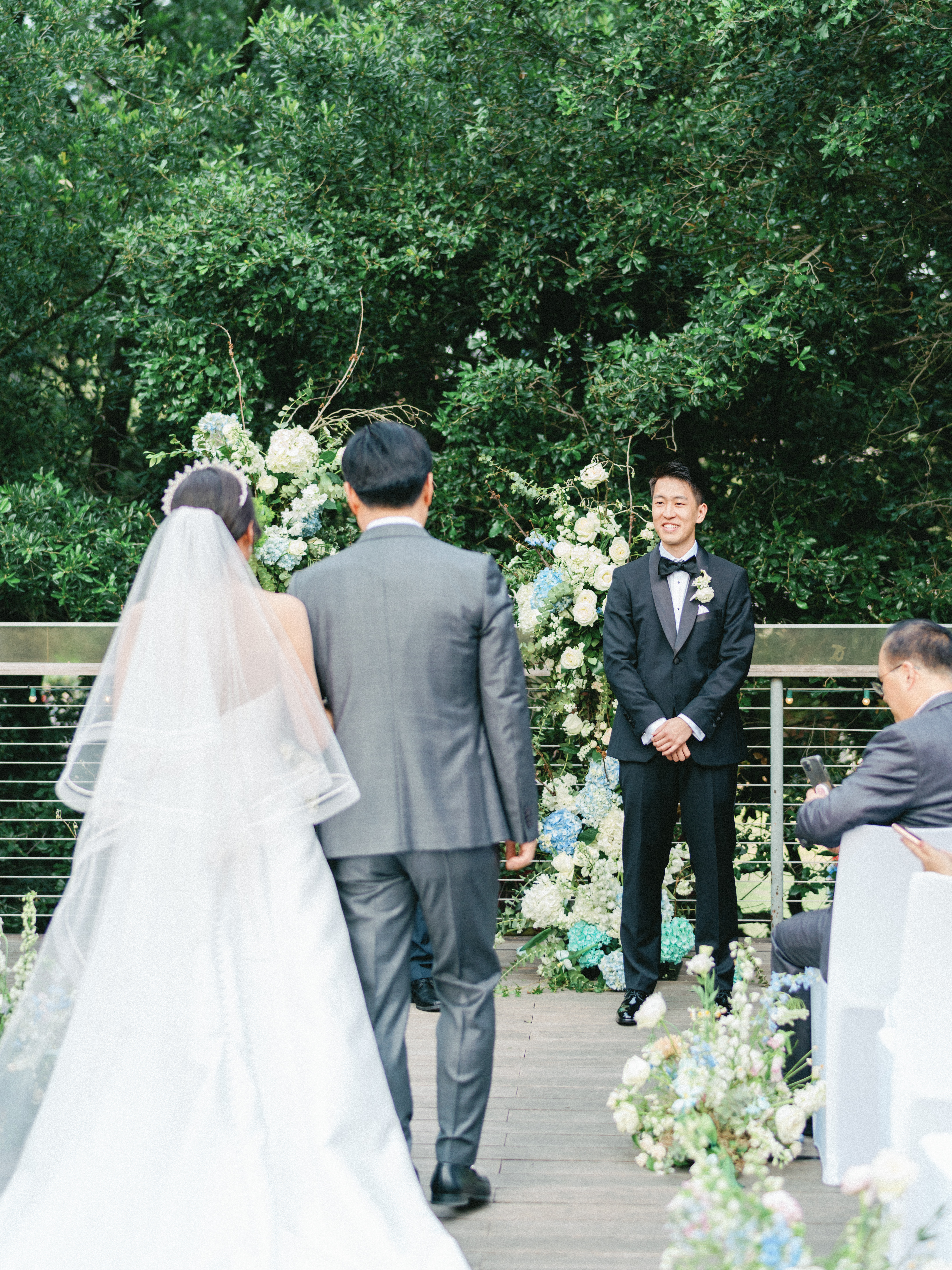 A wedding processional toward a floral arch accented with white roses, blue hydrangeas, and lush garden greenery.
