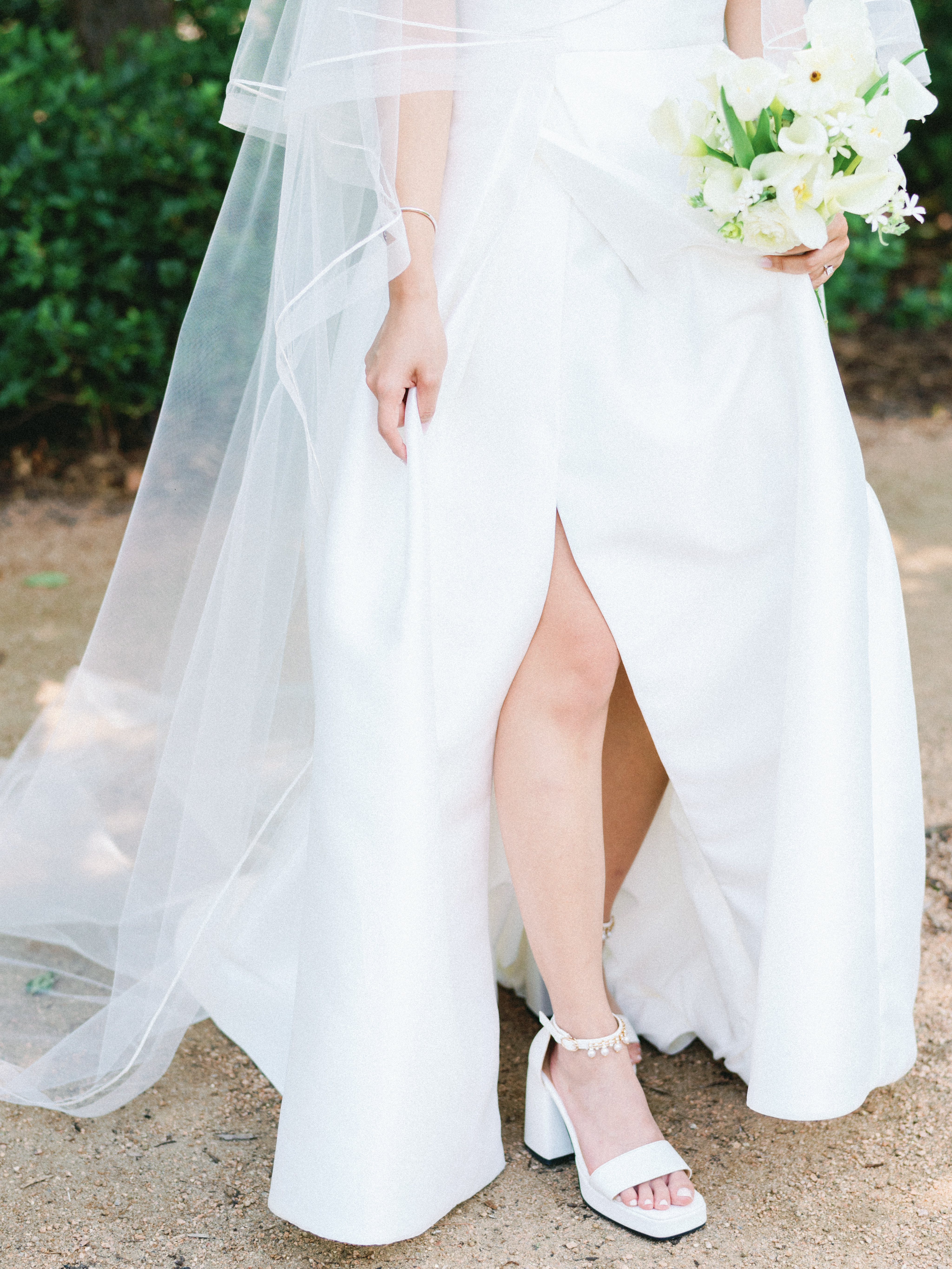 A bride lifts a white gown with a high slit, revealing white platform heels adorned with pearls along the ankle strap.