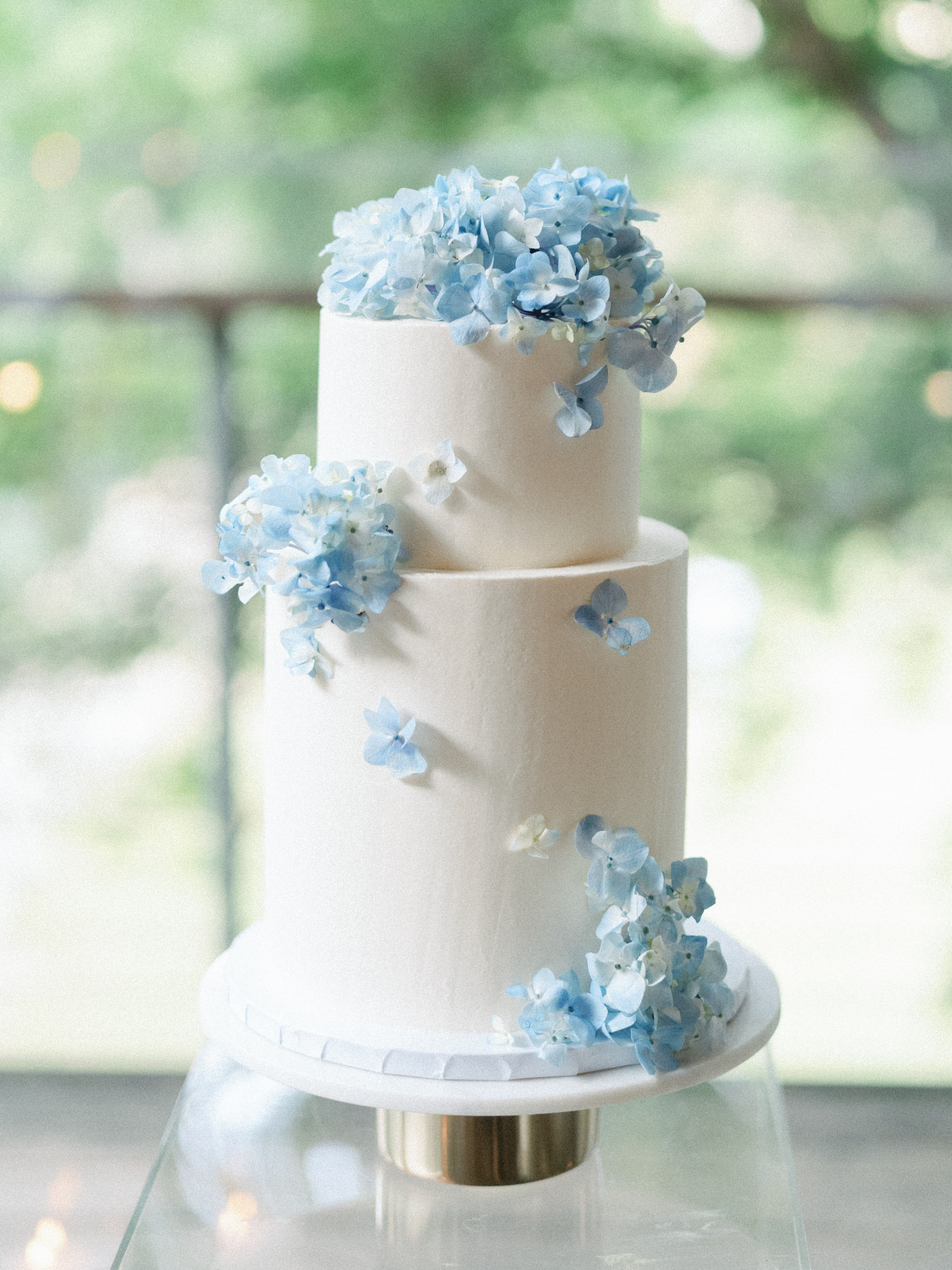 A smooth tiered white wedding cake decorated with delicate light blue hydrangea blossoms on a modern cake stand.